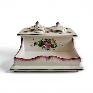 May include: A white ceramic desk organizer featuring a central floral motif with pink, green, and yellow petals. The piece includes two lidded circular containers on the top tier and a wide, open storage tray at the base with a scalloped pink trim.