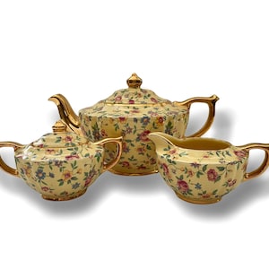 Könnte beinhalten: Ein Vintage-Tee-Set mit cremefarbener Basis und floralem Muster. Das Set enthält eine Teekanne, eine Milchkännchen und eine Zuckerdose. Die Teekanne hat einen goldenen Griff und Ausguss, und die Milchkännchen und Zuckerdose haben goldene Akzente.