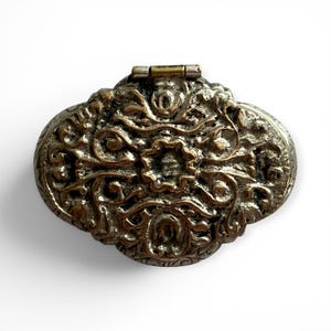 May include: An oval-shaped metal trinket box with a hinged lid. The exterior features a raised, textured relief pattern of swirling floral and scroll motifs in a tarnished silver and bronze finish, centered around a prominent flower design.
