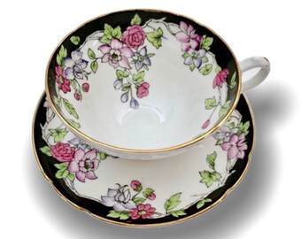 May include: A white teacup and saucer set with a black rim and gold trim. The cup and saucer are decorated with pink, purple, and green floral patterns. The teacup has a small handle.