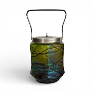May include: A vintage green glass jar with a silver lid and handle. The jar has a unique, textured design with a mix of green, blue, and gold hues. The handle is a simple, curved metal bar.