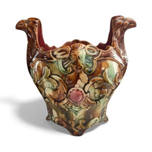 May include: A brown ceramic planter with a detailed, ornate design. The planter features two bird-like handles and is decorated with floral and geometric patterns in shades of brown, green, and red. The planter has a wide opening and a textured surface.