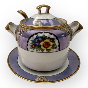 May include: A vintage porcelain sugar bowl with a lid and a small spoon, resting on a matching saucer. The bowl and saucer are white with lavender and gold accents. A floral design decorates the front of the bowl.