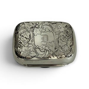 May include: A rectangular silver box with intricate leaf and vine engravings. The box features a central shield with the letter "D" and is likely made of sterling silver. The design is detailed and ornate, with a polished finish.