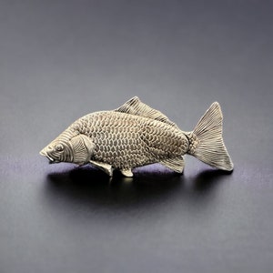 May include: A silver-colored fish figurine with detailed scales and fins. The fish is lying on its side with its head turned slightly to the left.
