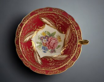May include: A red teacup with a gold rim and floral design. The cup has a white center with a floral pattern and a red rim with gold accents.