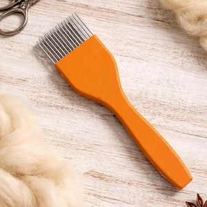 May include: An orange wool comb with metal tines, used for fiber preparation, lies on a white wooden surface.  Scissors, wool roving, buttons, and star anise are also present, suggesting a crafting theme.