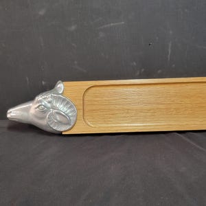 May include: A rectangular wooden tray with a recessed center. A silver-colored ram's head adorns one end. The tray is made of light-colored wood and is suitable for serving or display. The ram's head is detailed with horns and facial features.