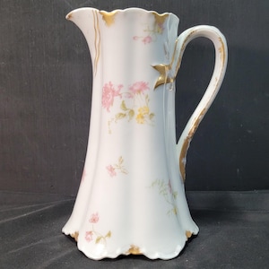 以下が含まれることがあります： 白磁のピッチャー。フレアベースとカーブしたハンドルが付いています。ピンクと黄色の花の模様と金のトリムで装飾されています。リムとハンドルも金でアクセントがつけられています。ピッチャーはスカラップエッジです。