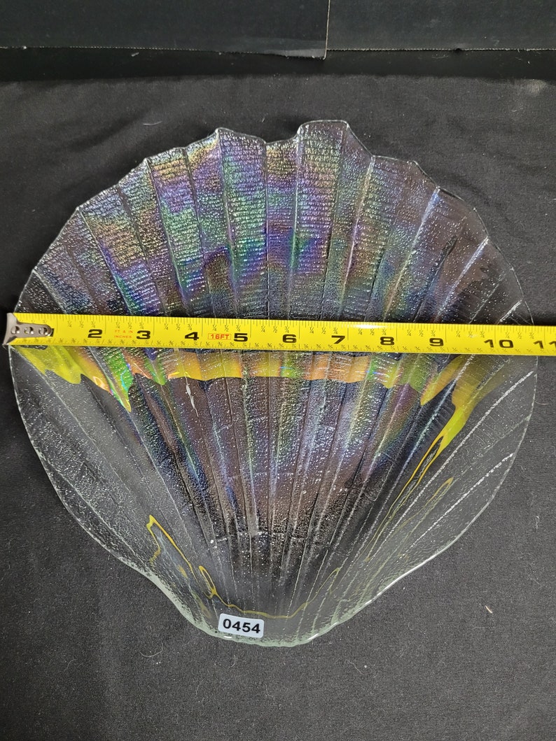 May include: A clear glass iridescent seashell-shaped dish with a textured surface. The dish is approximately 10 inches wide. The number 0454 is written on the bottom of the dish.