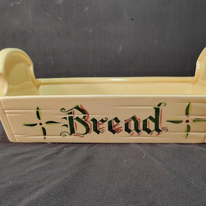 May include: Cream-colored ceramic bread basket with two handles. The front of the basket features the word "Bread" in a decorative font, with green and red accents. The basket has a rectangular shape with a textured, wood-like appearance.