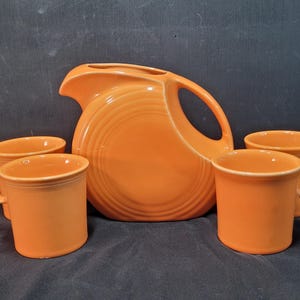May include: A bright orange ceramic pitcher with a circular body and a curved handle, accompanied by four matching orange ceramic mugs. Each piece features a smooth, solid-colored finish and a simple, functional design for serving beverages.