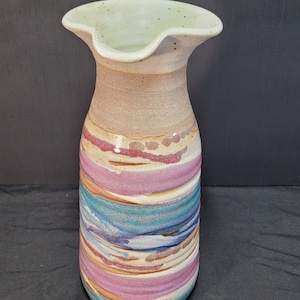 Puede incluir: Un jarrón de cerámica con una abertura ancha y acampanada y una base estrecha. El jarrón está decorado con un patrón de rayas coloridas en tonos de rosa, azul y marrón.