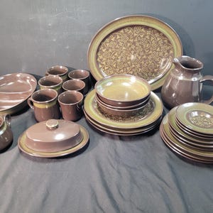 May include: A collection of brown and green ceramic tableware, including plates, bowls, cups, a serving platter, a jug, a creamer, and a butter dish. The set features a floral design and a two-tone colour scheme.