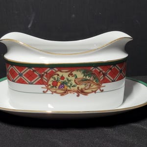 May include: White gravy boat with a matching saucer. The gravy boat has a gold trim and a decorative band with a red plaid pattern and a central illustration of fruit and a bird. The saucer has a green border.