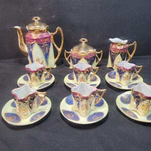 May include: A vintage tea set comprising a teapot, sugar bowl, creamer, and six cups and saucers. The set has gold trim, a cream background, and blue and burgundy accents. The teapot features a gold lid and handle.