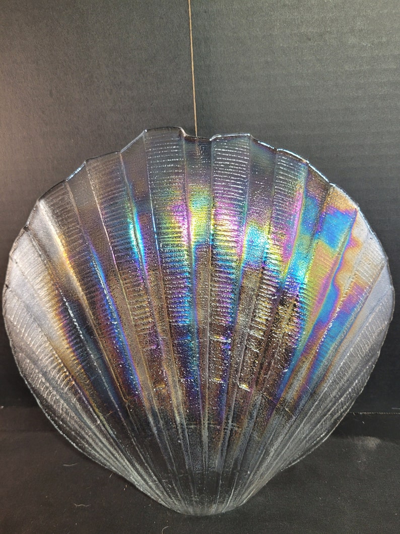 May include: A iridescent glass seashell dish with a textured surface. The dish is shaped like a scallop shell and has a light blue, purple, and green iridescent finish.