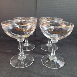 Op de afbeelding: Set van vier heldere champagne coupe glazen. Elk glas heeft een gouden tarwe ontwerp op de kom. De glazen hebben een steel en een brede, ondiepe kom, perfect voor het serveren van champagne of cocktails.