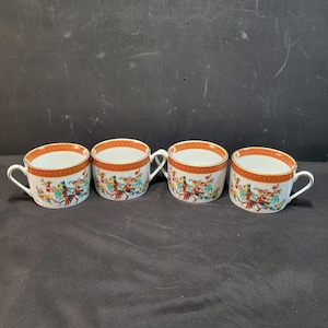 May include: Four white teacups with handles, featuring a vibrant design of figures and floral patterns in red, blue, green, and yellow. The rims have a red and gold geometric border. The cups are arranged in a row.
