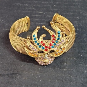 May include: Gold-toned metal cuff bracelet featuring a decorative mask design. The mask is adorned with colorful rhinestones in shades of red, blue, orange, and purple. The bracelet has a mesh-like texture.