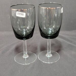 May include: A pair of stemmed wine glasses. The bowls are dark grey with silver rims, and the stems and bases are clear. Each glass has a small white sticker with the number "0996".