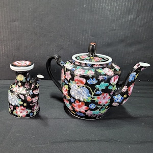 Könnte beinhalten: Ein schwarzes Keramik-Teeservice mit einem Blumendesign in Rot, Rosa, Blau und Grün. Die Teekanne hat einen schwarzen Griff und Ausguss. Der Sahnekännchen ist ein kleiner, passender Krug.