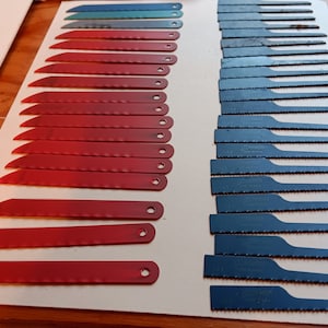 May include: A close-up of a set of saw blades for a jigsaw. The blades are arranged in two rows, with the red blades on the left and the blue blades on the right. The blades are all the same size and shape, but they have different teeth patterns.