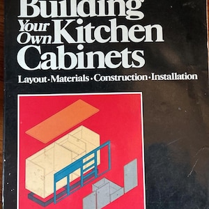 May include: A book cover with the title "Building Your Own Kitchen Cabinets" in white letters on a black background. The cover features an illustration of cabinet components and the author's name, Jere Cary.