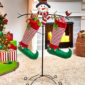 Puede incluir: Un soporte para calcetines navideños con dos calcetines a rayas rojas y blancas con zapatos de elfo verdes. Una cabeza de muñeco de nieve y un pequeño oso de peluche decoran el soporte. Un árbol de Navidad y regalos están en el fondo.