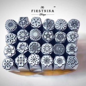 Könnte beinhalten: Eine Sammlung von vierundzwanzig Metallstempeln, die in einem Raster angeordnet sind und jeweils ein einzigartiges Blumenmuster auf der Vorderseite aufweisen. Der Text "The FIRSTNINA Design" steht über den Stempeln. Die Stempel sind silberfarben.