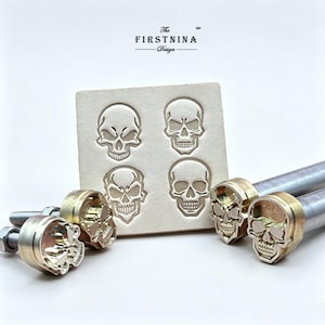 May include: A set of four metal skull stamps with threaded bolts and two metal handles, accompanied by a square piece of light-colored leather featuring four embossed skull impressions. The text "The FIRSTNINA Design" is visible at the top.
