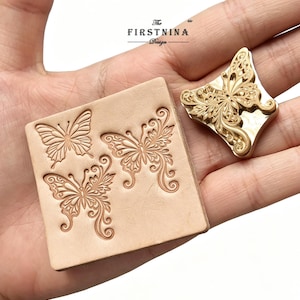 May include: A square piece of light tan leather featuring two embossed butterfly designs and a separate gold-toned metal butterfly stamp. The stamp has intricate filigree details and the text The FIRSTNINA Design is visible above.