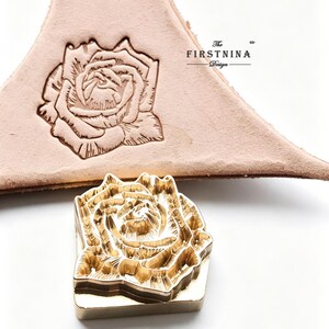 May include: A gold-colored metal stamp featuring a detailed rose design, positioned below a piece of light tan leather embossed with the same rose pattern. The text "The FIRSTNINA Design" is printed in black on the leather surface.