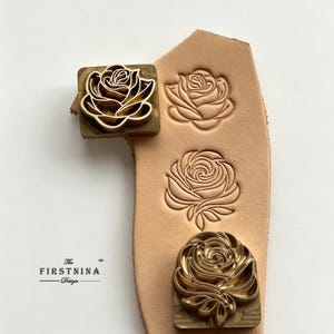 May include: A brass metal stamp featuring a rose design, positioned on a piece of light tan leather. The leather displays two distinct rose impressions created by the stamp. The text "The FIRSTNINA Design" is printed in black on the left side.
