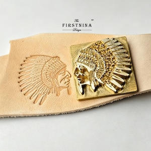 May include: A square brass leather stamping tool featuring a detailed relief of a person wearing a feathered headdress. The tool is positioned next to a matching impression stamped into a piece of light tan leather. The text THE FIRSTNINA Design is visible.