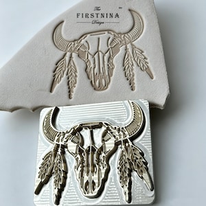 May include: A square metal stamping tool featuring a bull skull design with feathers, positioned below a piece of light beige leather embossed with the same bull skull and feathers, and the text The FIRSTNINA Design printed above the skull.