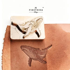 May include: A rectangular brass metal stamp featuring a raised humpback whale design, positioned above a piece of tan leather embossed with the same whale image. The text "The FIRSTNINA Design" is printed in black above the metal stamp.