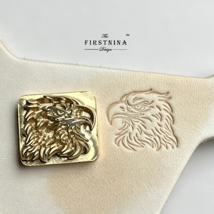 May include: A square brass metal stamp featuring a detailed eagle head design, positioned next to an identical eagle head impression embossed into a piece of light-colored leather. The text FIRSTNINA Design is printed above the stamp.