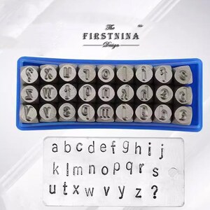 May include: A set of metal alphabet stamps arranged in a blue plastic tray, featuring individual letters and a question mark. Below the tray, a rectangular metal plate displays the stamped lowercase alphabet and a question mark in black ink.