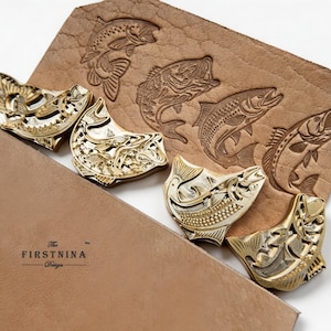 May include: Four gold-toned metal fish-shaped stamps arranged on a piece of tan leather. The leather features four embossed fish designs matching the stamps. The text "The FIRSTNINA Design" is printed in black on the lower left corner of the leather.