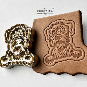May include: A gold-toned metal stamp featuring a detailed engraving of a scruffy dog face with paws, positioned next to a piece of tan leather embossed with the same dog design. The text "The FIRSTNINA Design" is printed above the stamp and leather.