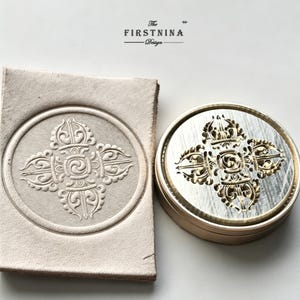 May include: A circular metal stamp featuring an intricate four-pronged vajra design and a matching embossed impression on a square piece of light beige leather. The text "The FIRSTNINA Design" is printed in black above the stamp and leather piece.