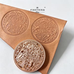 May include: A light brown leather sheet featuring two circular embossed designs of an Egyptian deity with a jackal head, alongside a matching circular metal stamp tool. The text "THE FIRSTNINA Design" is printed in black above the leather.