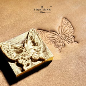 May include: A brass metal stamp featuring a butterfly design, positioned next to a matching butterfly impression on a tan leather surface. The stamp is engraved with the text The FIRSTNINA Design, which is also visible in the leather imprint.