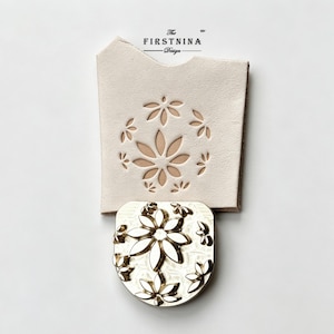 May include: A light beige leather piece featuring a laser-cut floral pattern and the text The FIRSTNINA Design, paired with a matching metallic silver-toned stamp tool displaying an identical embossed flower motif for leather crafting.