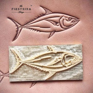 May include: A rectangular metal leather stamping tool featuring a raised tuna fish design. The stamp is positioned over a piece of tan leather that shows a clear, debossed impression of the same tuna fish, with the text The FIRSTNINA Design.