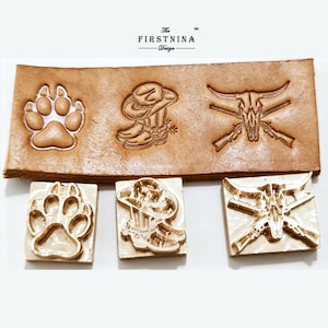 May include: Three brass metal stamps featuring a paw print, a cowboy boot with a hat, and a steer skull with crossed rifles. Below the stamps is a tan leather strip showing the embossed impressions of these three distinct western designs.