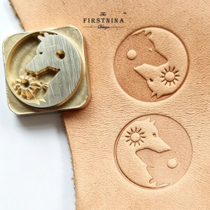 May include: A square brass leather stamping tool featuring a wolf head design with a sun motif, positioned next to two circular impressions on a piece of tan leather. The stamp is engraved with the text THE FIRSTNINA Design.