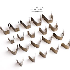 May include: A collection of twenty-one bronze-toned metal leather craft cutting dies arranged in a grid. Each die features a V-shaped or curved blade edge. The text "The FIRSTNINA Design" is printed in black at the top center of the image.