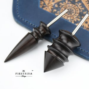 May include: Two dark brown wooden burnishing tools for leathercraft, each featuring a smooth, contoured handle with a pointed tip and a protruding metal rod. The text "The FIRSTNINA Design" is printed in black at the bottom left of the image.